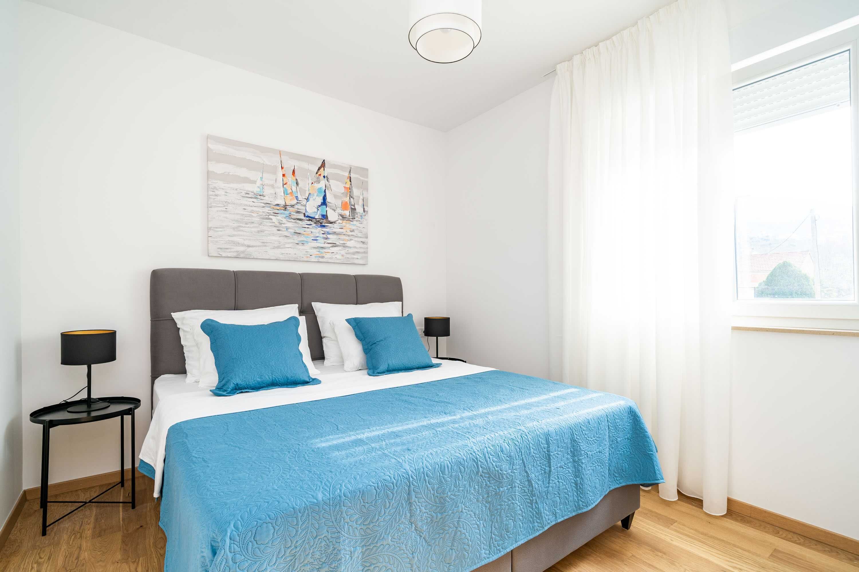 Una cama bien hecha con colcha azul, dos almohadas a juego y un cabecero. En la pared, un cuadro con barcos a vela, una ventana con cortinas blancas y una mesita de noche con una lámpara al lado de la cama.