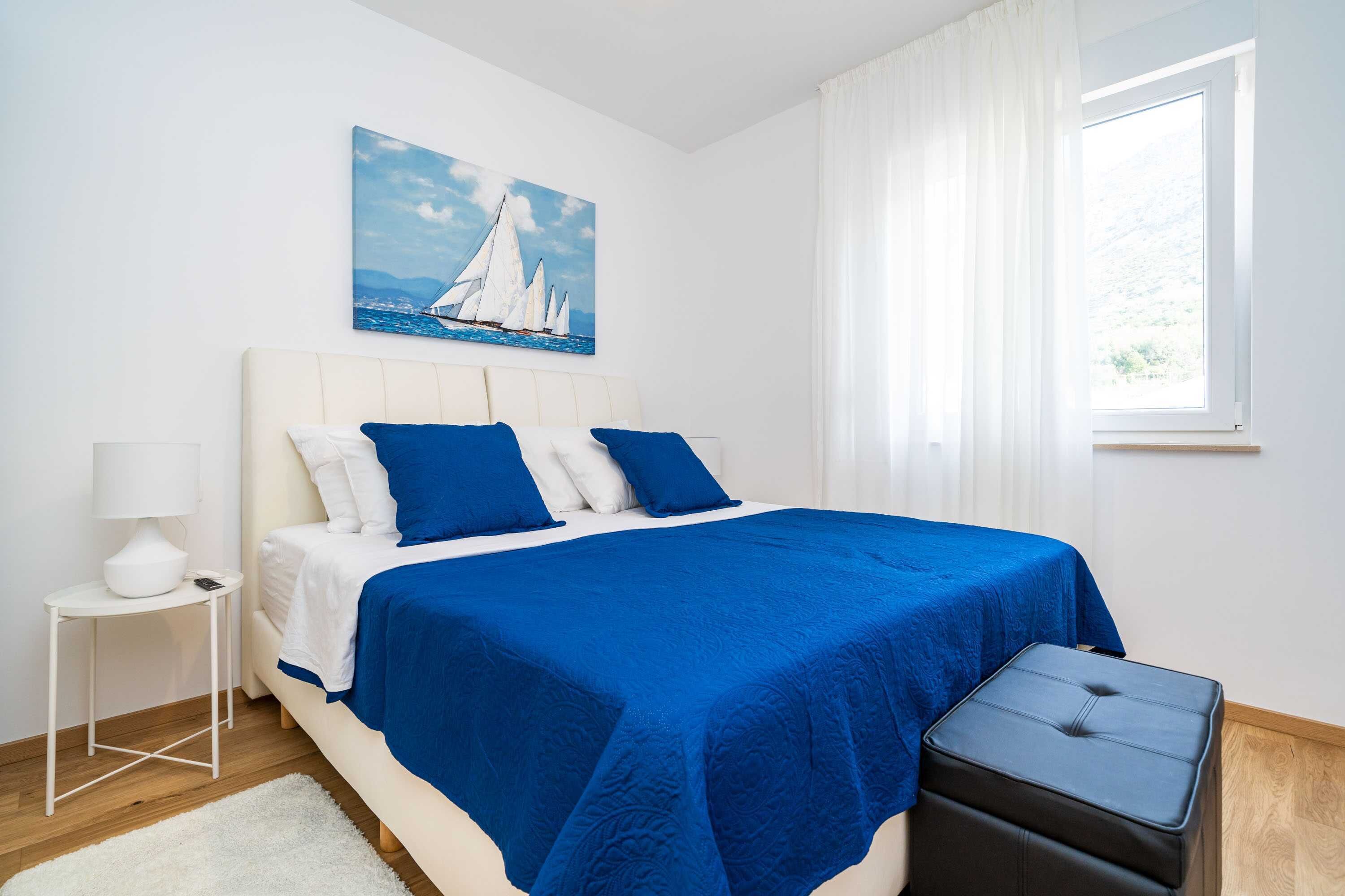 A bedroom with a bed, a nightstand, a window, and a painting of sailboats.