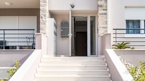 Una entrada de edificio moderna con una escalera que lleva a la puerta, una cámara de seguridad encima de la puerta y un pequeño espacio de estar al aire libre a la izquierda.