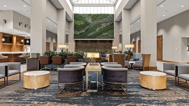 A spacious lobby with a high ceiling, a central seating area with round tables and chairs, and a large green wall at the back. - Embassy Suites by Hilton Irving Las Colinas (Irving)