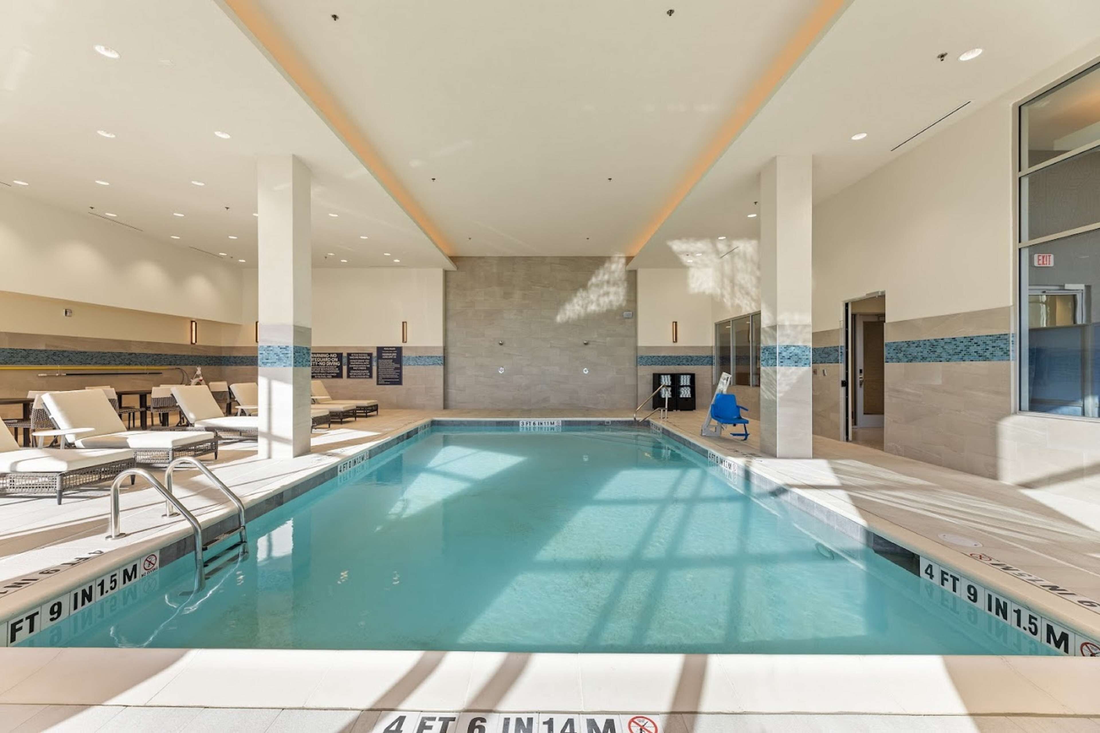 Una piscina interior con agua cristalina, rodeada de sillas de descanso y un suelo de baldosas.