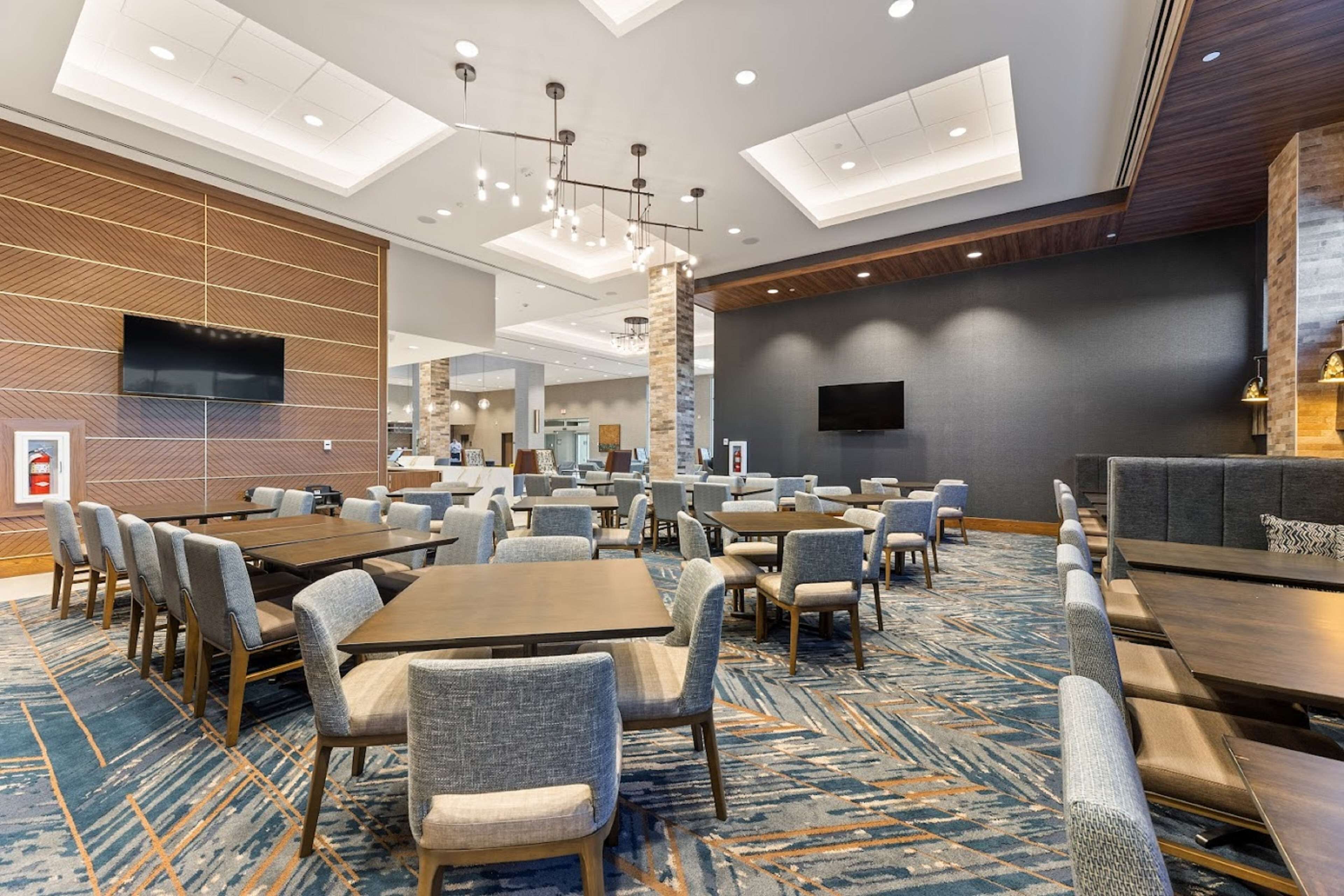 A hotel lobby with multiple seating areas, including tables and chairs, a large flat-screen TV mounted on the wall, and a patterned carpet.