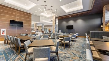 A hotel lobby with multiple seating areas, including tables and chairs, a large flat-screen TV mounted on the wall, and a patterned carpet.