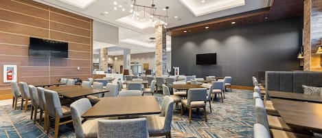 A hotel lobby with multiple seating areas, including tables and chairs, a large flat-screen TV mounted on the wall, and a patterned carpet.