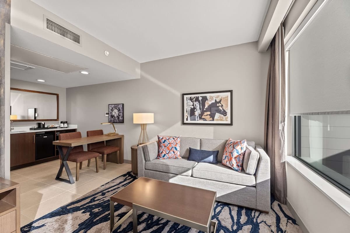 A modern hotel room with a dining area, a sofa set, a coffee table, and a large window.