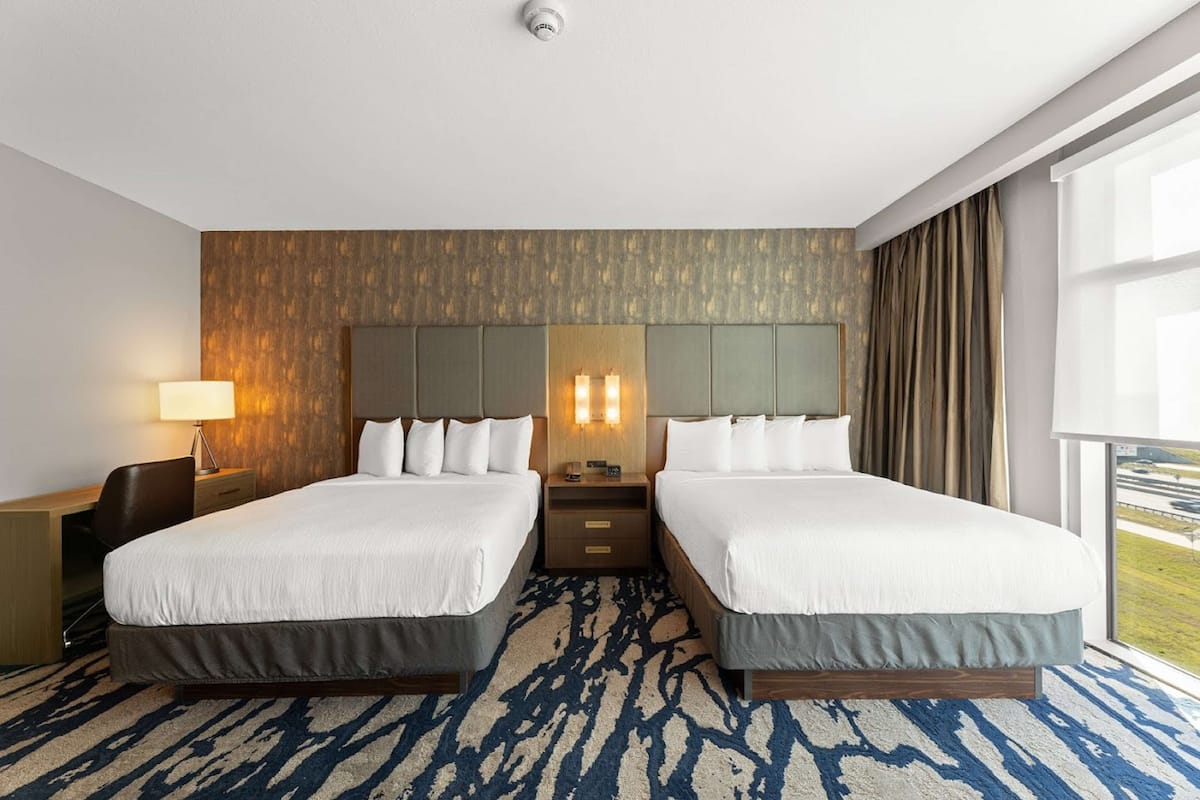 A hotel room with two beds, a desk, a lamp, and a view of the outdoors.