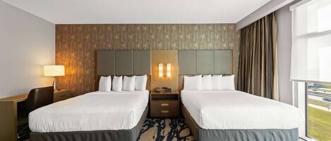 A hotel room with two beds, a desk, a lamp, and a view of the outdoors.