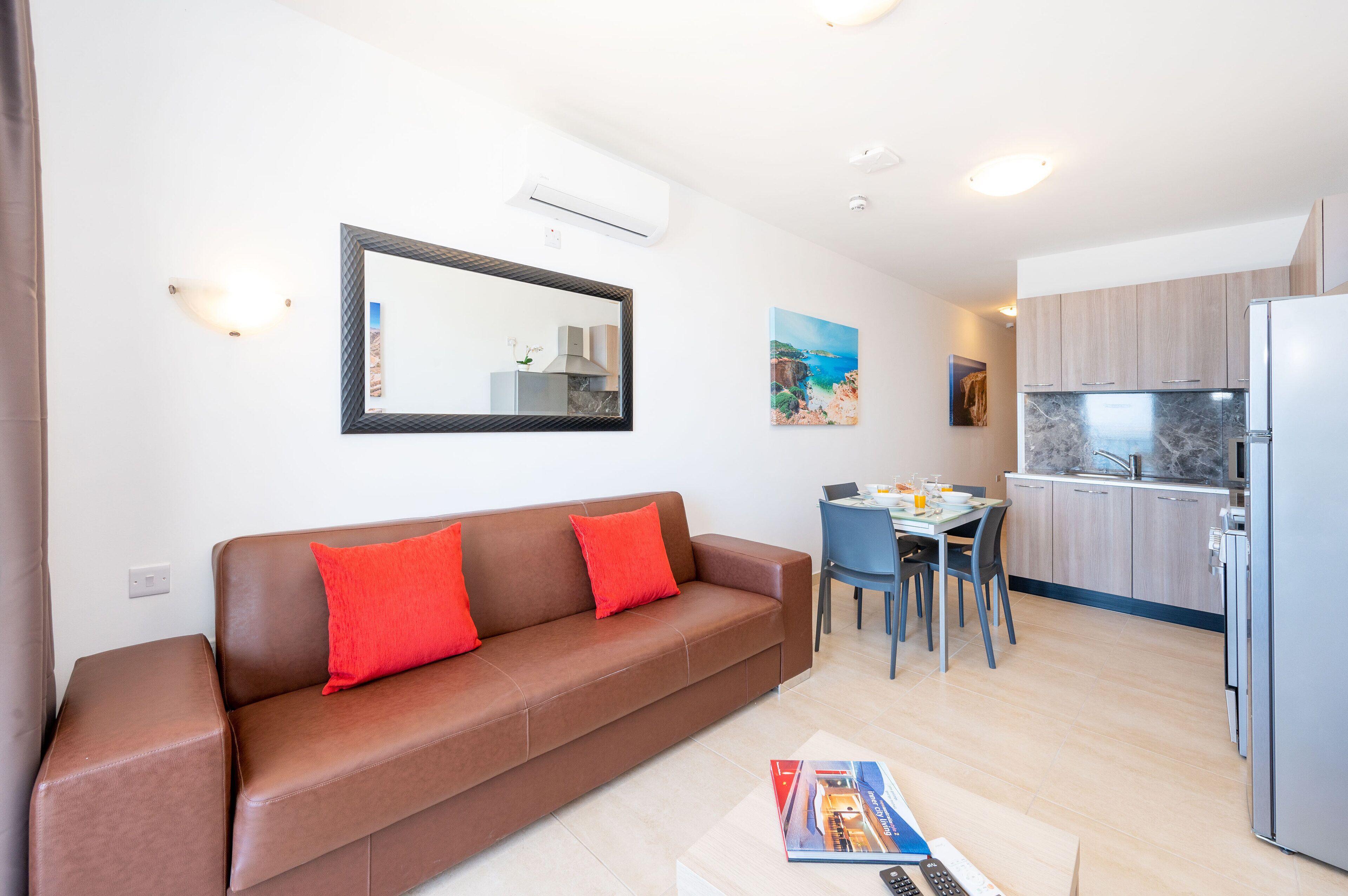 A modern living room with a brown leather sofa, a dining table with chairs, a kitchen area with a refrigerator, and a large mirror on the wall.