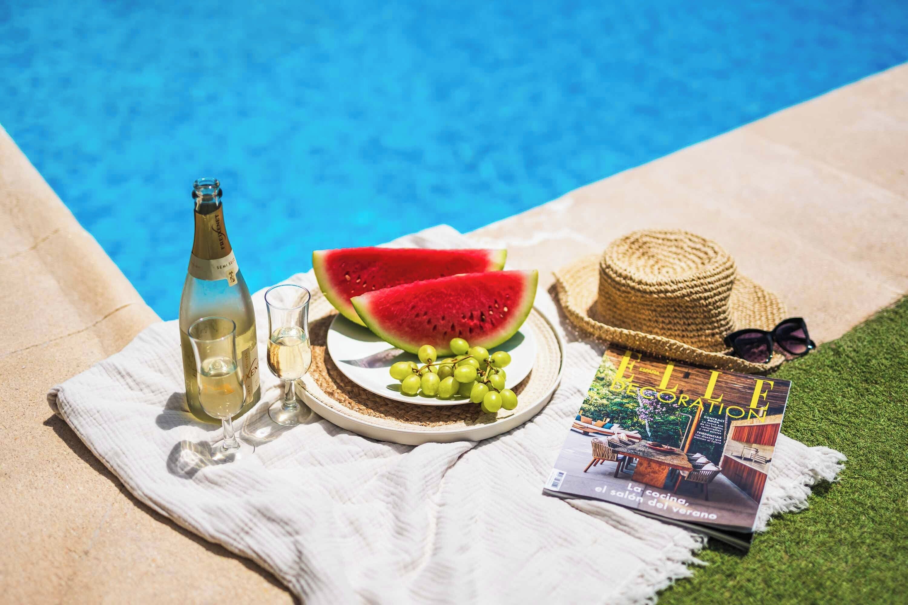A bottle of white wine, two glasses, a plate with watermelon and grapes, a straw hat, and a magazine on a towel by a pool.