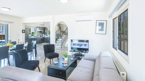 A modern living room with a sofa, a coffee table, and a TV. There is a dining area with chairs and a table. - Villa Delfini 16 - Four Bedroom Villa, Sleeps 8 (Protaras)