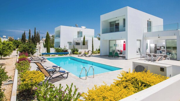 A modern pool area with a clear blue pool, lounge chairs, and a white building with large windows. - Protaras Greco Villa CCV2 (Πρωταράς)