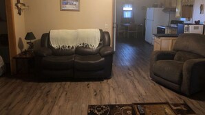 Ein Wohnzimmer mit einem dunkelbraunen Ledersofa, einem kleinen Tisch mit einer Lampe und Holzboden. Im Hintergrund ist eine Küche mit Kühlschrank und Mikrowelle zu sehen.