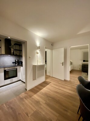 A modern kitchen with built-in appliances, a dining area with a black chair, and a hallway leading to a bedroom.