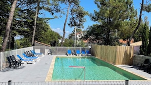 Une piscine entourée d’arbres et d’une zone délimitée par une clôture, avec des chaises longues à proximité.