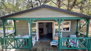 Une petite cabane en bois avec un porche couvert, deux chaises en plastique blanc, une table et un banc.