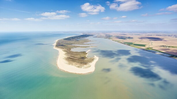 Vue aérienne d’une zone côtière avec une plage de sable, des eaux cristallines et, au loin, des terres agricoles et des bâtiments.