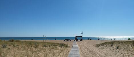 A sandy beach with a wooden boardwalk leading to the water, a flag, and people in the distance.