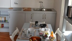 A kitchen with white cabinets, a sink, and a dining table with chairs. There are shelves above the cabinets holding various kitchen items.