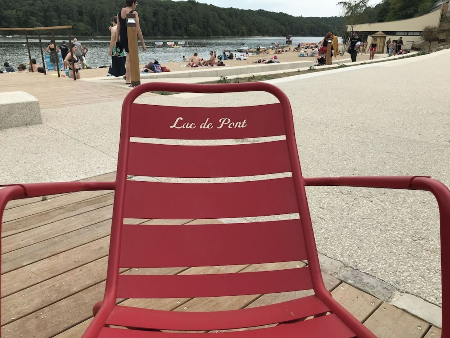 Una sedia rossa con la scritta "Lac de Pont", un molo di legno e una spiaggia sullo sfondo con persone.