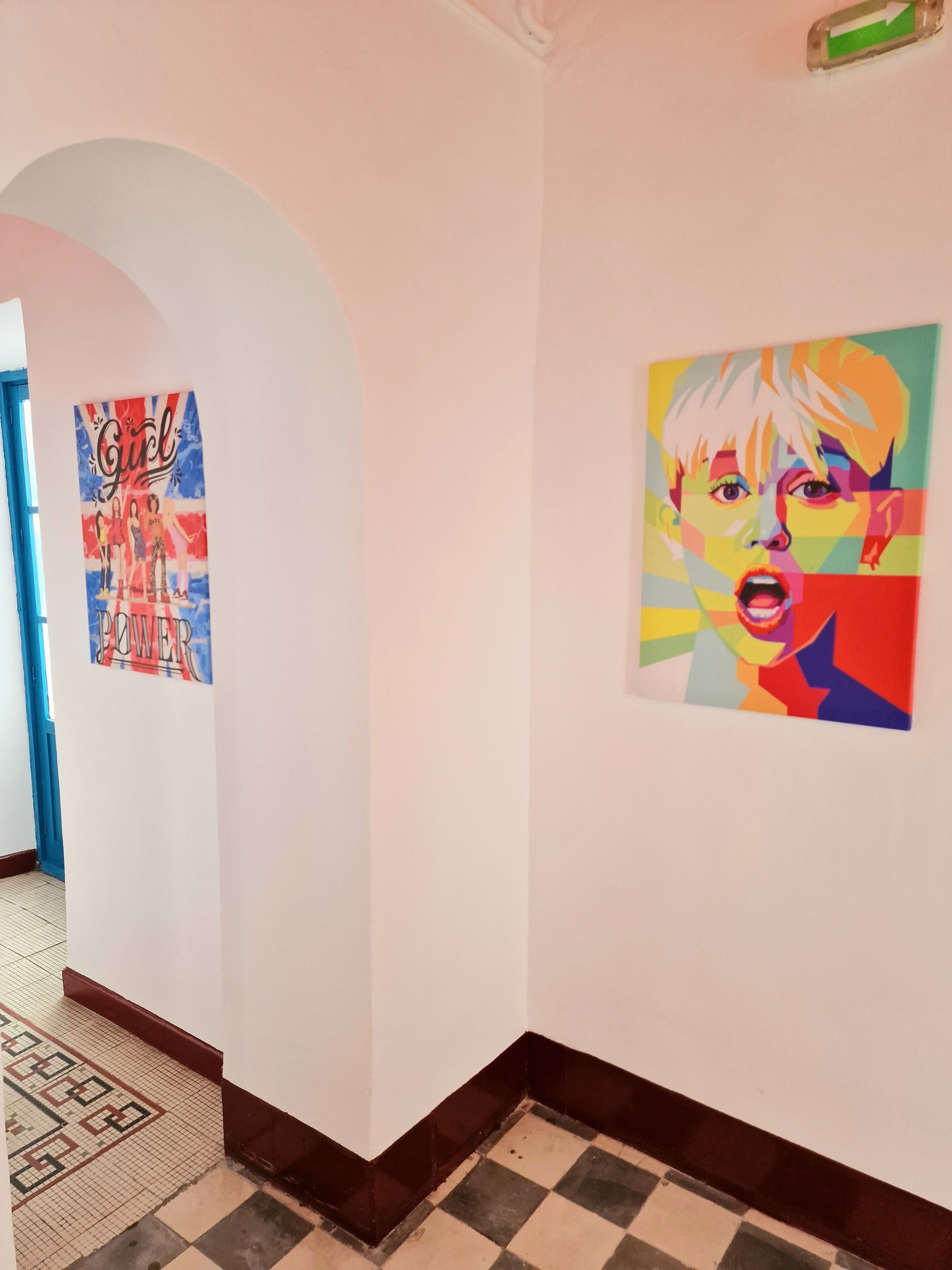Un couloir doté d’un sol carrelé, une affiche accrochée au mur de gauche et un tableau coloré au mur de droite.