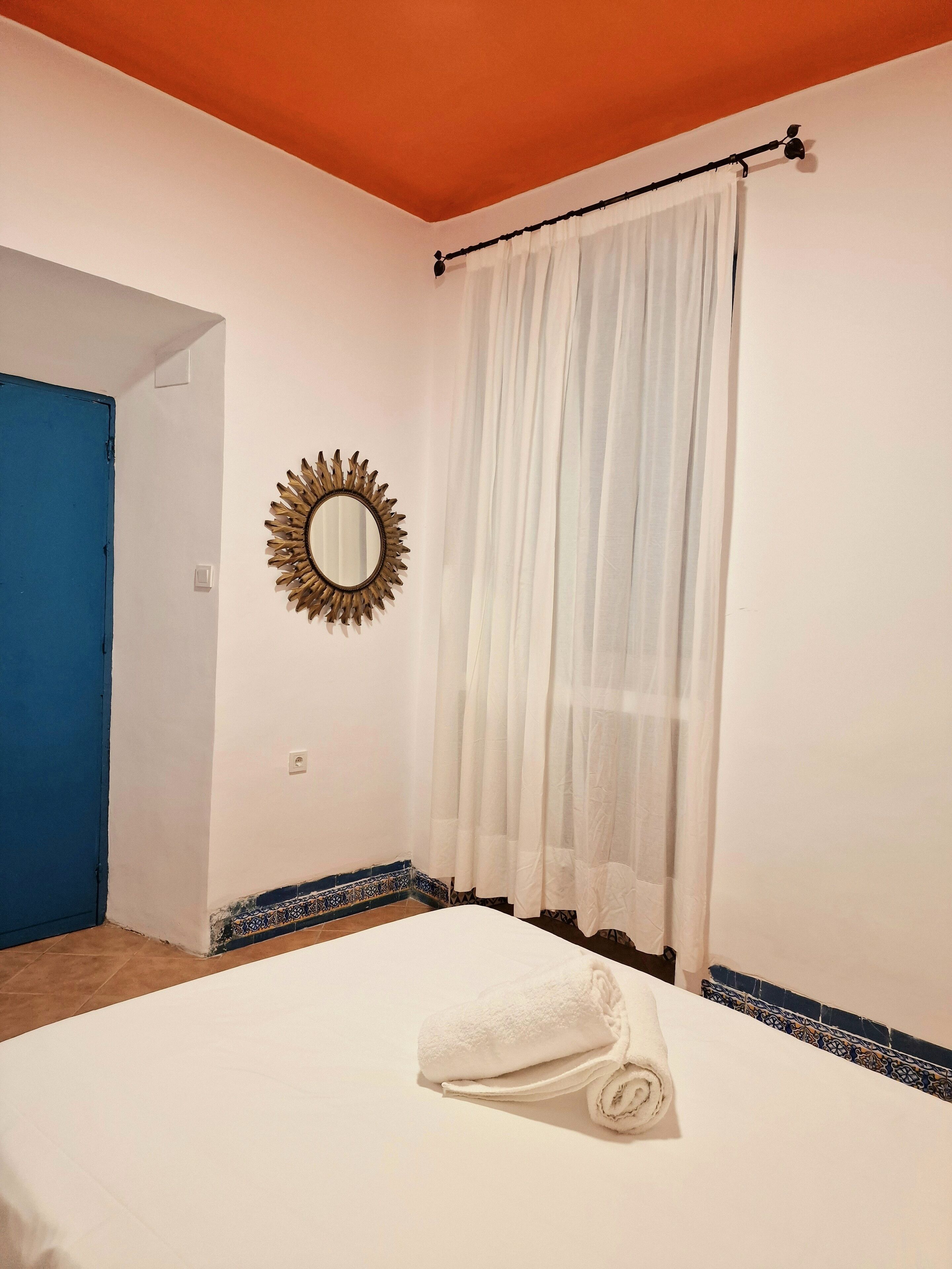 Una habitación de hotel con una cama, una cortina blanca, un espejo con forma de sol y una puerta azul.