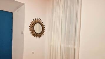 Una habitación de hotel con una cama, una cortina blanca, un espejo con forma de sol y una puerta azul.