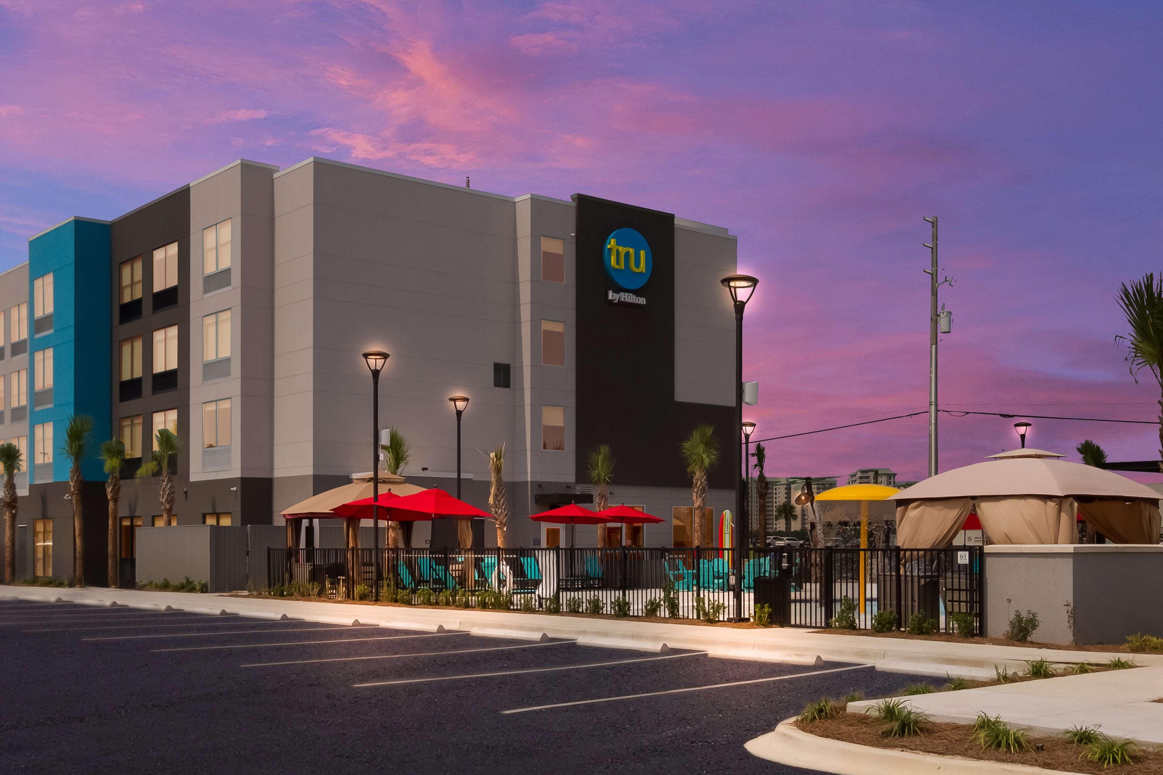 Un hotel modern amb un cartell ben visible de 'tru by Hilton', ombrelles exteriors i un aparcament.
