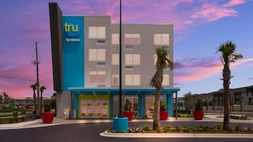 A modern hotel with a blue and white facade, named 'tru by Hilton', featuring a central entrance, large windows, and surrounding landscaping.
