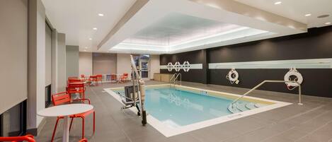 Zona de piscina con sillas y mesas rojas, un puesto de salvavidas y una pared oscura con un adorno blanco.