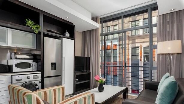 Un soggiorno moderno con un divano, poltrone a righe, un tavolino da caffè e una TV. È presente un angolo cottura con elettrodomestici e un forno a microonde. La stanza ha ampie finestre con tende e vista sugli edifici esterni.