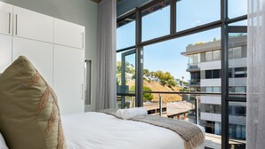 Una habitación de hotel moderna con una cama grande, vistas de la ciudad a través de ventanas de piso a techo y un balcón con vistas de los edificios circundantes.