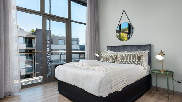 Un dormitorio moderno con una cama grande, un espejo redondo y vistas a un edificio a través de un ventanal.