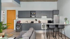 Una cocina moderna con gabinetes grises, lavadora integrada, microondas y refrigerador de acero inoxidable. Hay un sofá gris y una zona de bar con taburetes.