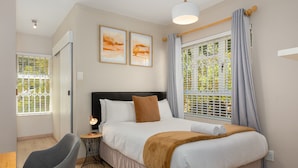 Een keurig opgemaakt slaapkamer met een bed, een nachtkastje met een lamp, een stoel en twee ingelijste kunstwerken aan de muur.