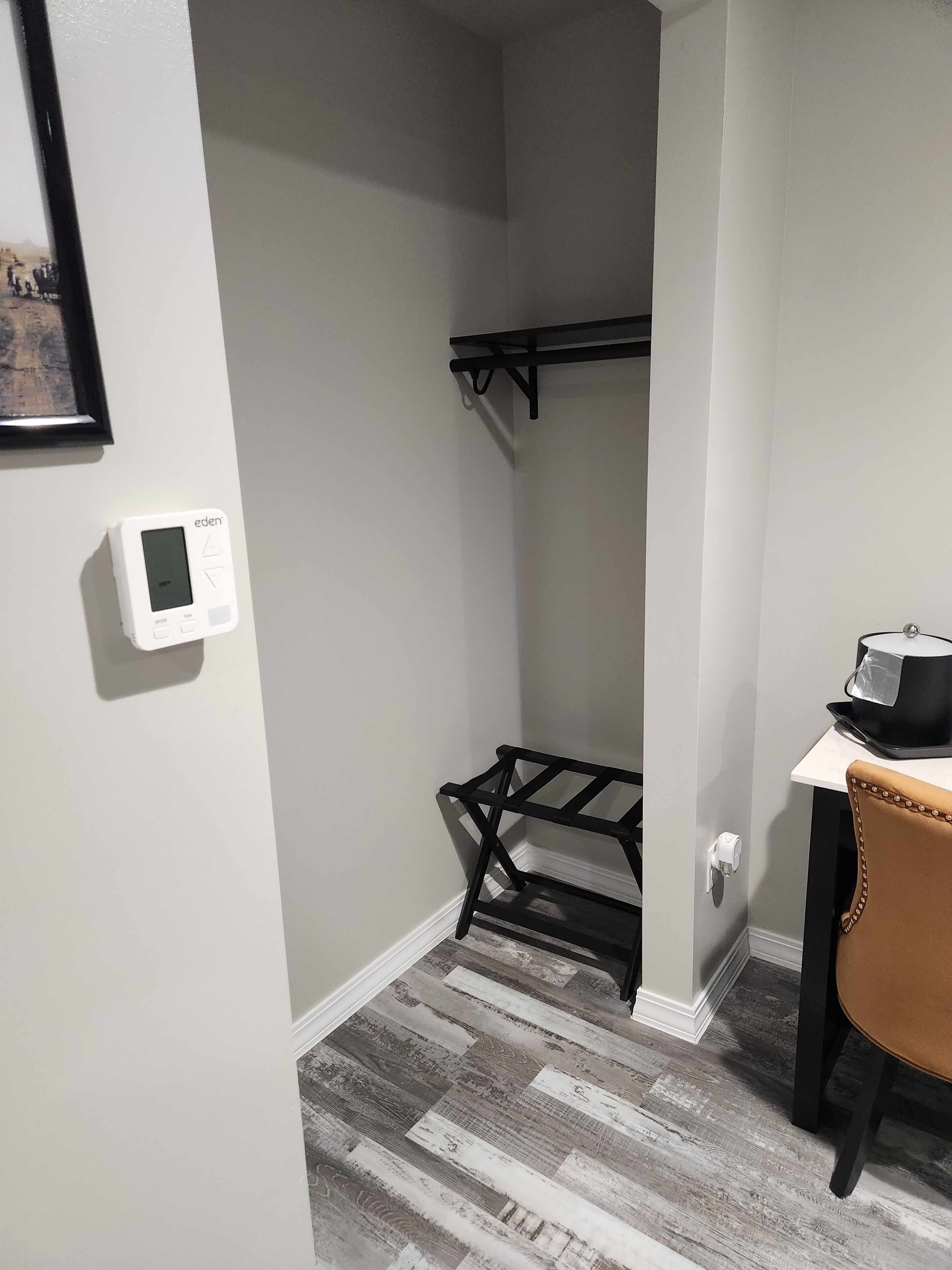 A small, narrow room with a white wall, a black metal shelf, a white thermostat, and a brown chair.