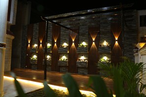 A modern outdoor patio area with a stone wall and wooden beams, illuminated by wall-mounted lights.
