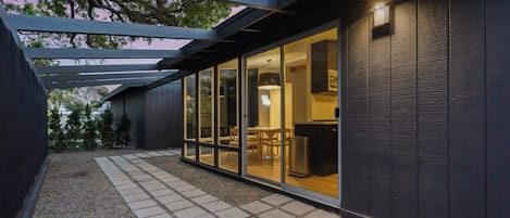 En moderne bygning med en overdækket terrasse, et spiseområde, der kan ses gennem store glasdøre, og en mørk facade.