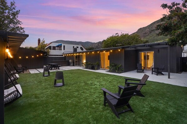 Un patio con césped artificial, mobiliario exterior e iluminación colgante.