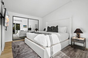 A modern bedroom with a large bed, a bedside table, a mirror, and a view of a balcony.