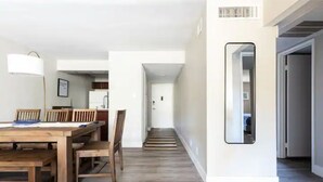 Een moderne keuken met witte kasten, een houten eettafel en stoelen. Een gang leidt naar een slaapkamer met een deur.