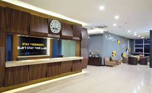 A hotel reception area with a wooden reception desk, a clock, and a wall with the text "ROMS INC." - Rooms Inc BTC Pasteur (Bandung)