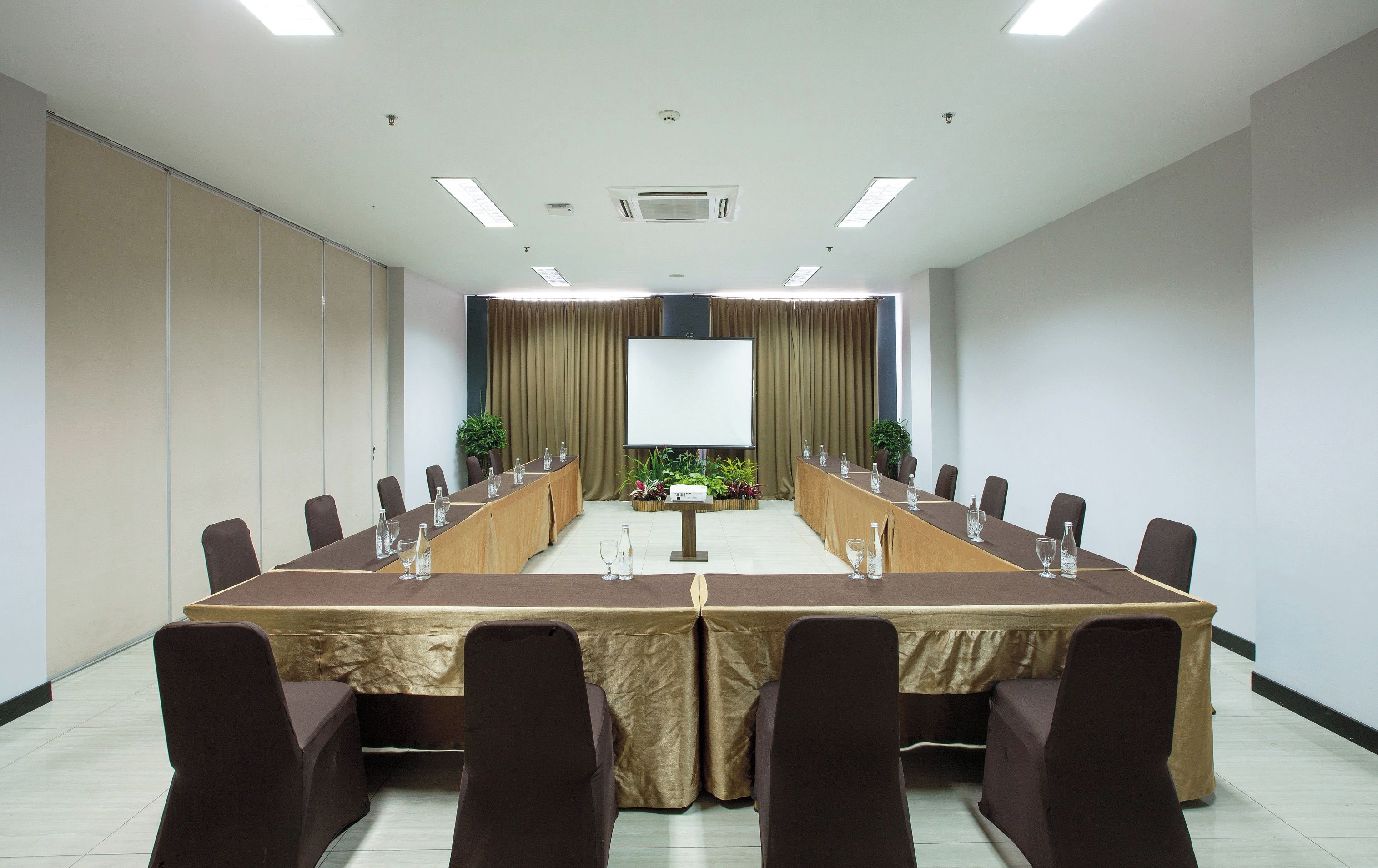 Uma sala de conferências com uma mesa comprida, cadeiras, uma tela de projetor e uma planta.
