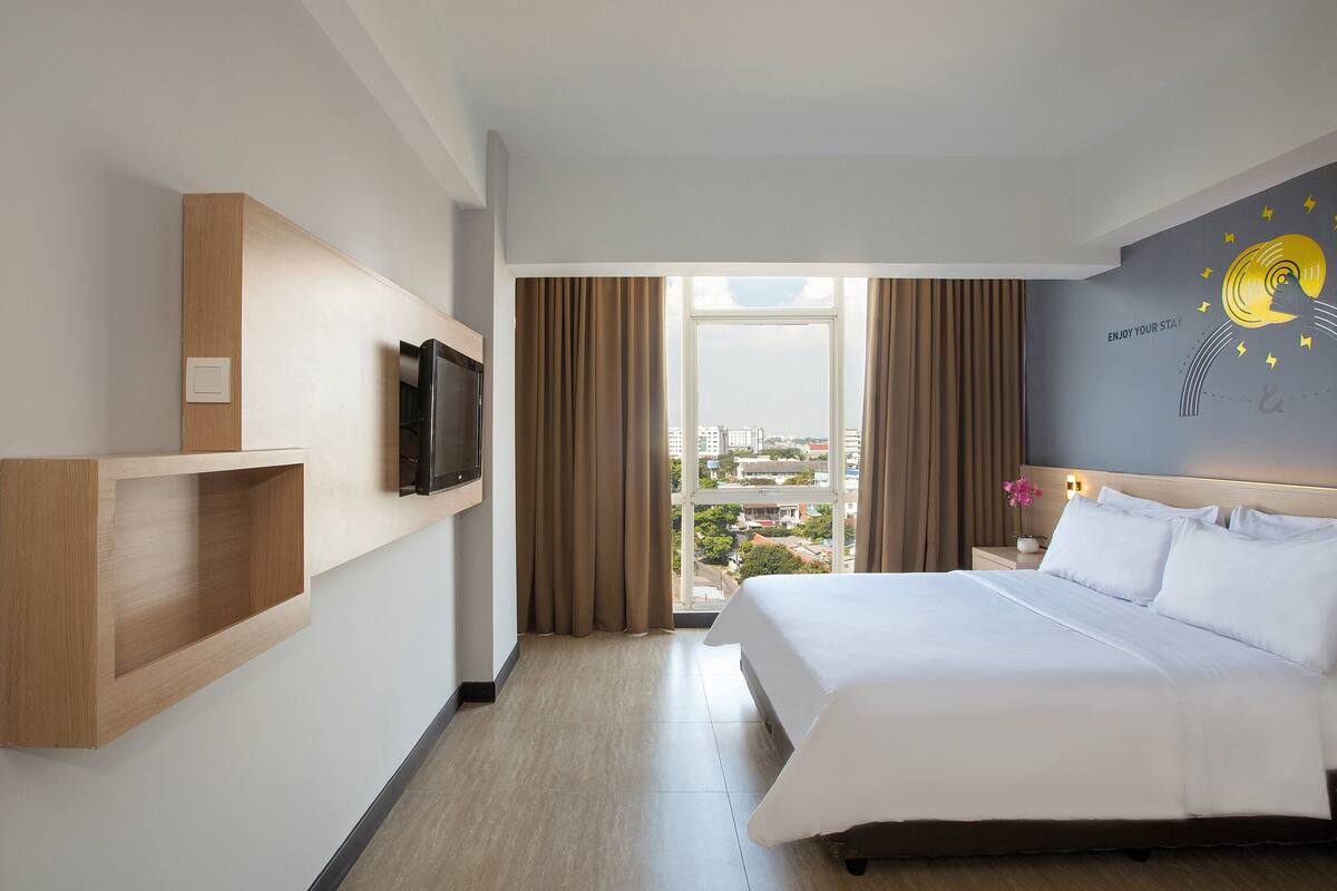 A hotel room with a large bed, a mounted TV, a wooden shelf, and a view of the city through the window.