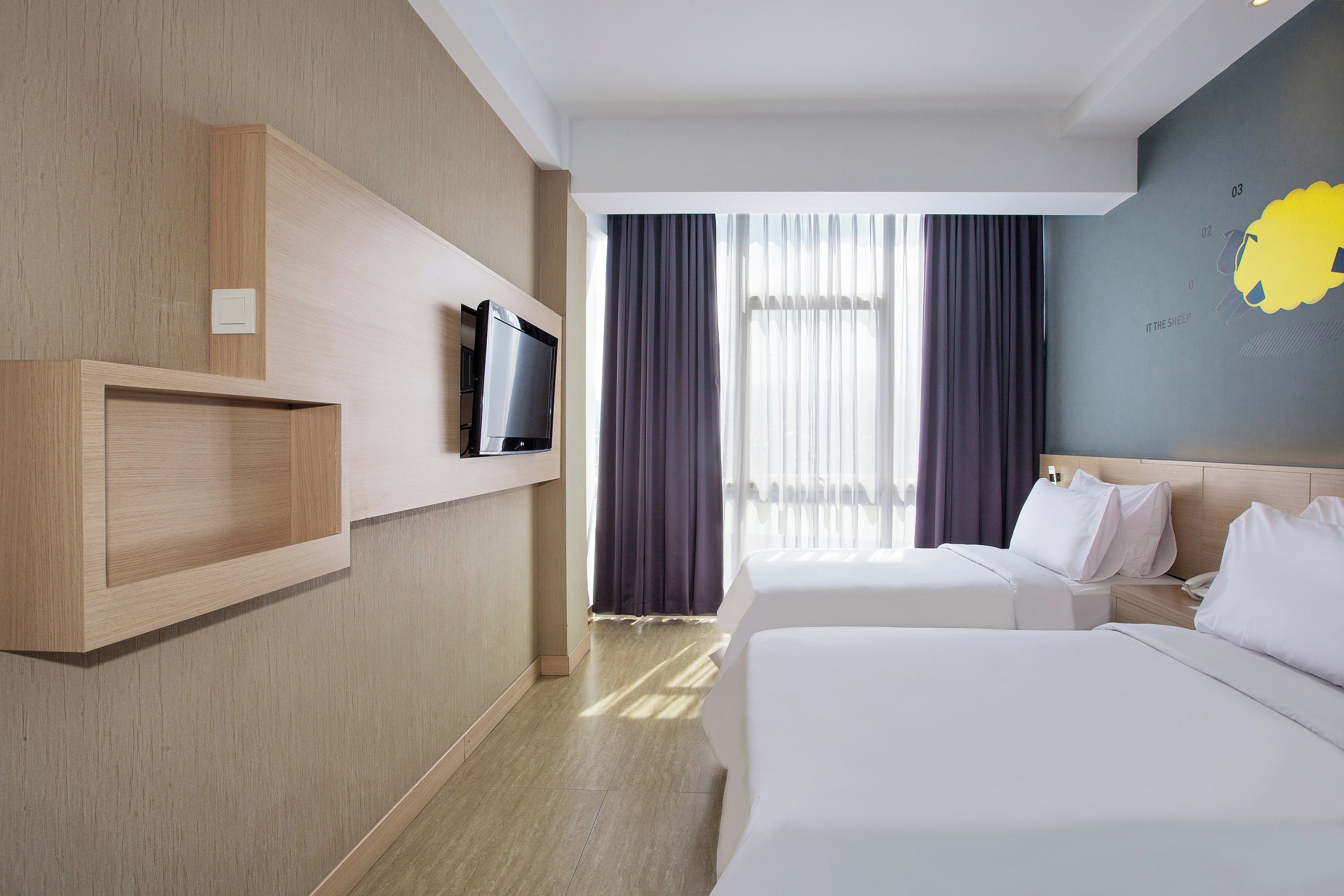 Ein Hotelzimmer mit zwei Betten, einem Fernseher auf einem Holzboden, einem Nachttisch und einem Fenster mit Vorhängen.