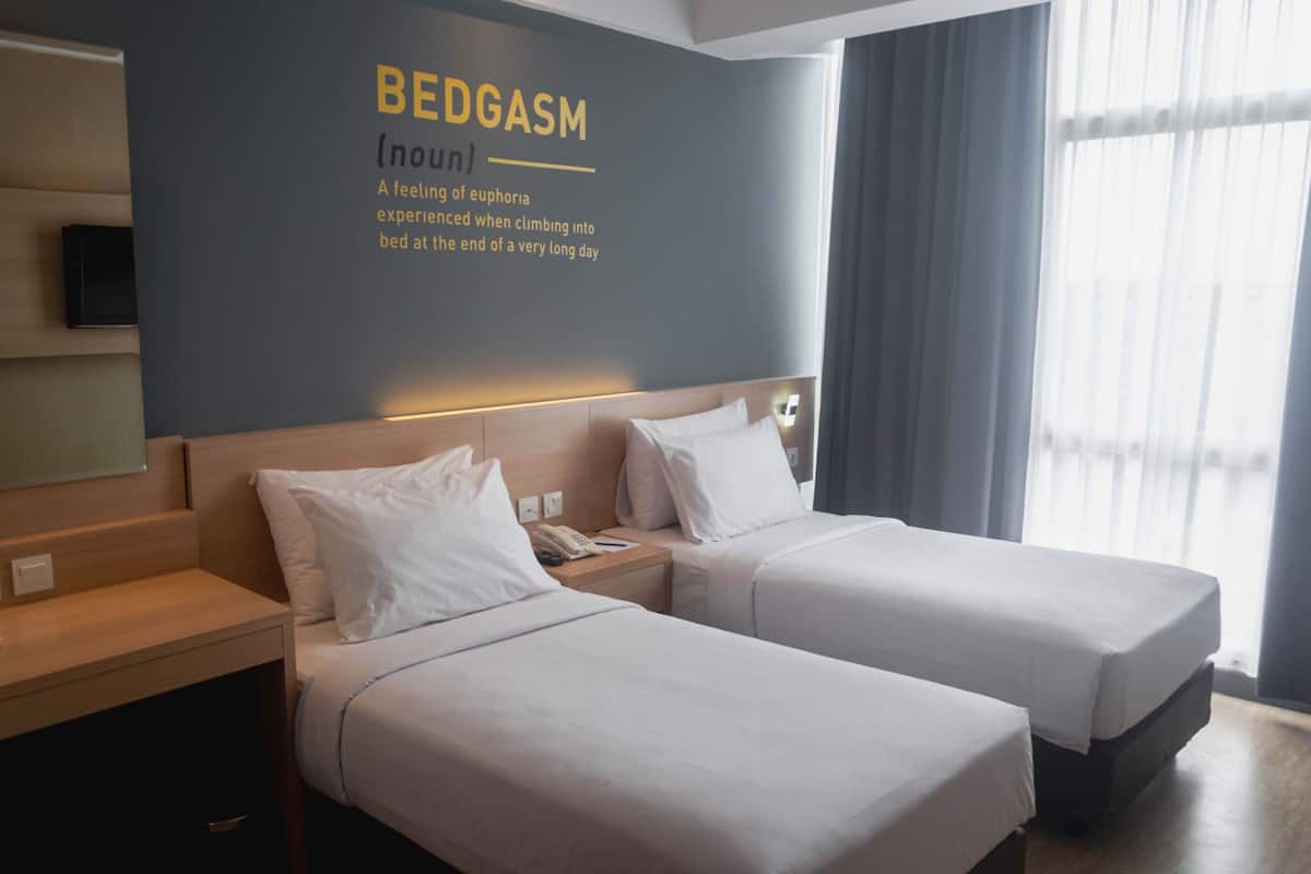 A hotel room with two beds, a wall-mounted TV, and a large window with curtains.