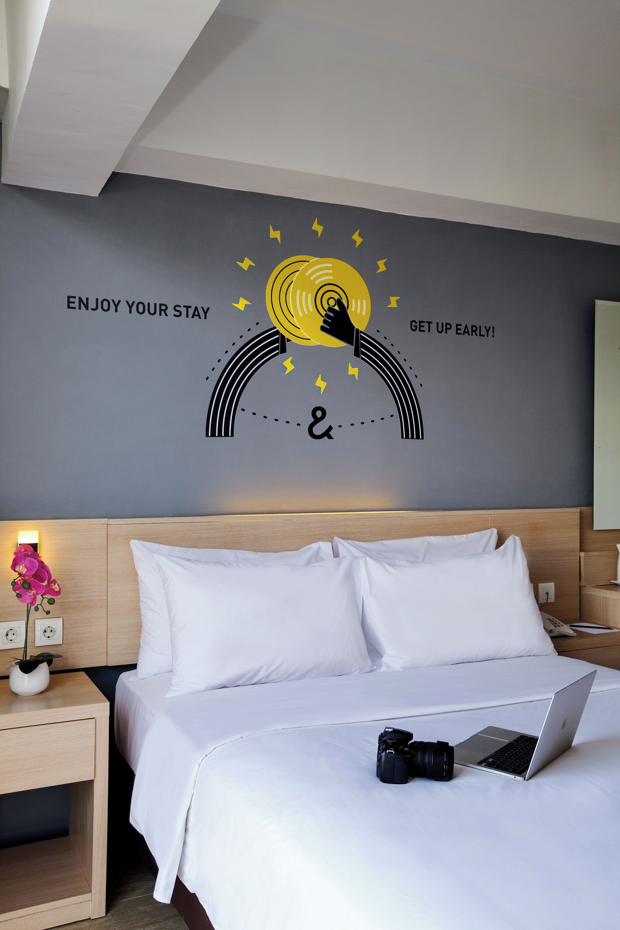 A hotel room with a bed, a nightstand with a lamp and a flower vase, and a wall with a motivational quote and a sun graphic.
