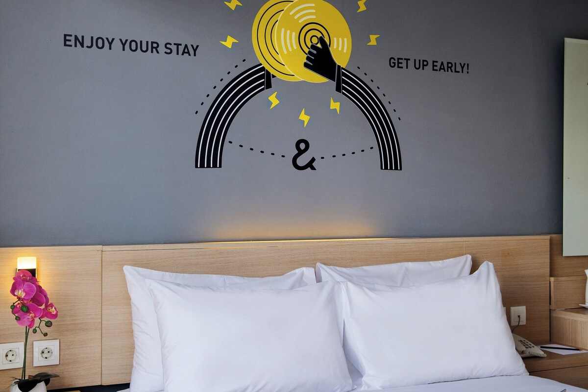 Hotelkamer met een bed, nachtkastje met een lamp en een bloevas, en een muur met een motiverende quote en een zonafbeelding.