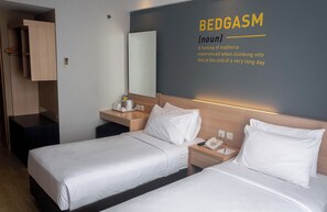 A hotel room with two beds, a bedside table with a phone and a lamp, a wall-mounted mirror, and a wall with a definition of 'BEDGASM'. - Rooms Inc BTC Pasteur (Bandung)