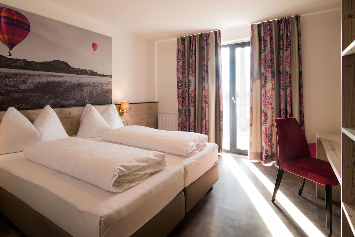 A hotel room with two beds, a red chair, a window with curtains, and a wall with a hot air balloon painting.