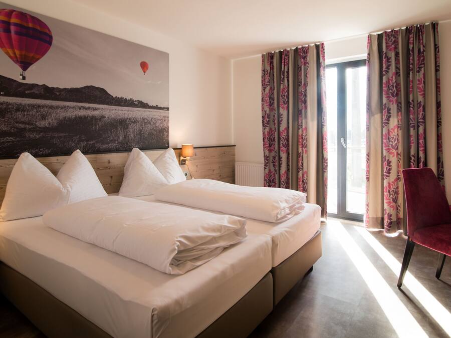 Habitación de hotel con dos camas, una silla roja, una ventana con cortinas y un cuadro de un globo aerostático en la pared.